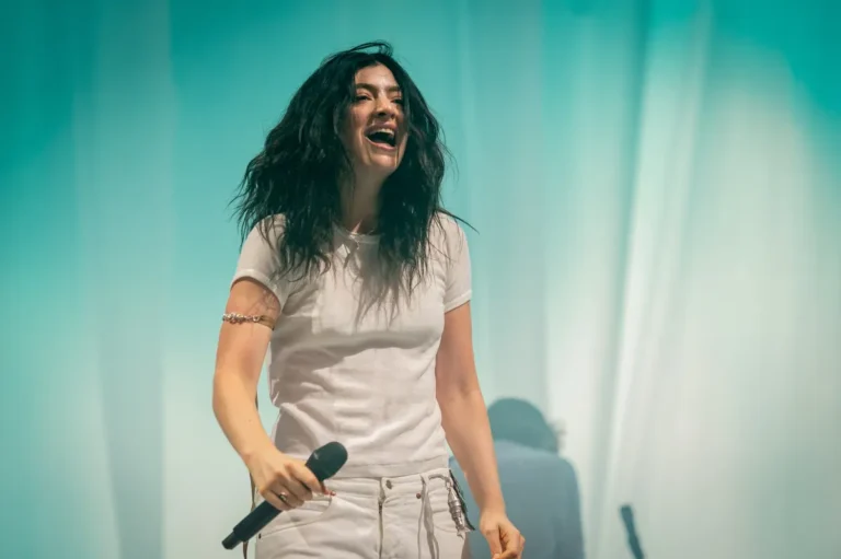 Person with long hair in a white outfit holding a microphone, smiling on stage with a teal backdrop.