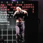 Performer on stage sings with a microphone, dark clothing, digital screen behind displays red and white text.