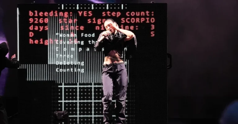 Performer on stage sings with a microphone, dark clothing, digital screen behind displays red and white text.