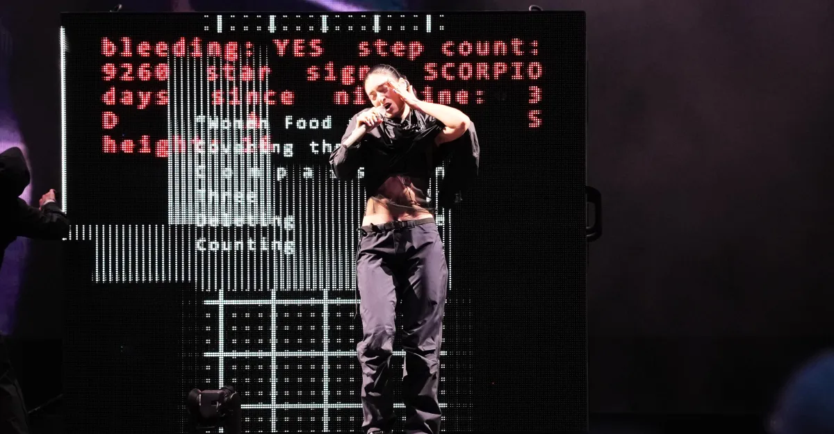 Performer on stage sings with a microphone, dark clothing, digital screen behind displays red and white text.