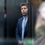 Man in a suit stands in an elevator, partially obscured by doors, looking outward with a neutral expression.