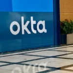 Okta logo on a blue wall with a reflective marble floor and a planter with green plants nearby.
