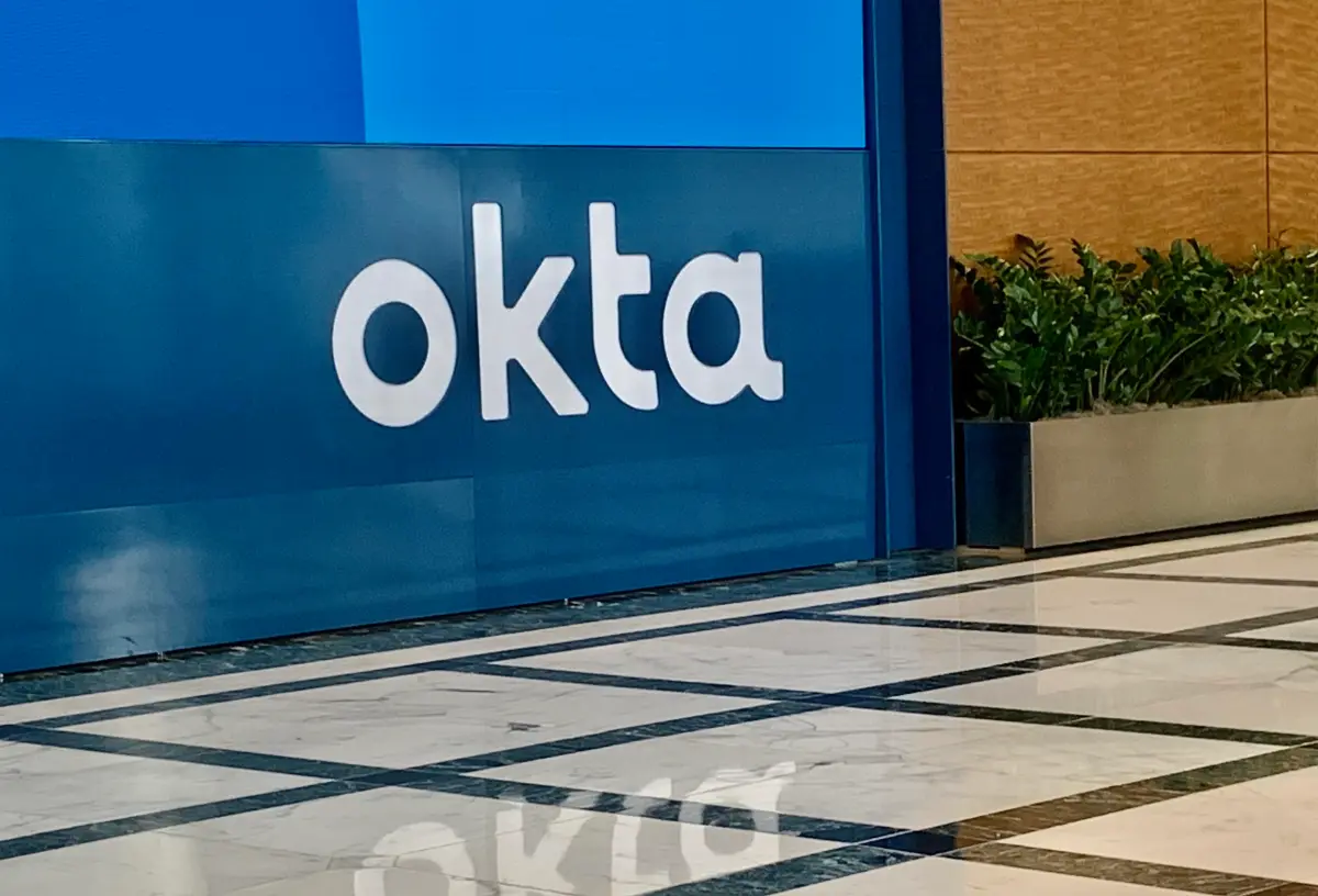 Okta logo on a blue wall with a reflective marble floor and a planter with green plants nearby.