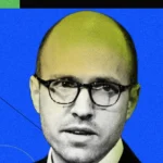 Man with glasses against a blue and green geometric background, wearing a suit and looking towards the camera.