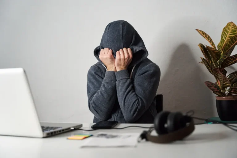 Person in a hoodie sitting at a desk with head in hands, laptop open, headphones, and a plant nearby.