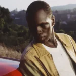 Person in a tan jacket leans against a red car with a small white device on the hood, set against a hilly landscape.