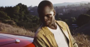Person in a tan jacket leans against a red car with a small white device on the hood, set against a hilly landscape.