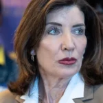 Woman with brown hair and white collar looks sideways; blurred individuals in the background, one with a denim jacket.