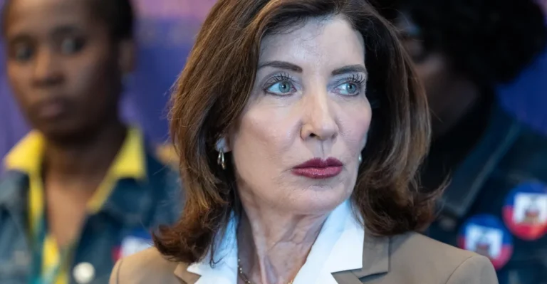 Woman with brown hair and white collar looks sideways; blurred individuals in the background, one with a denim jacket.