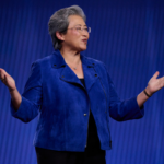 Person in a blue jacket speaking on stage, arms outstretched against a blue background.