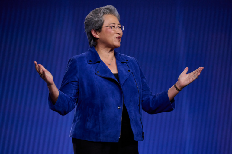 Person in a blue jacket speaking on stage, arms outstretched against a blue background.
