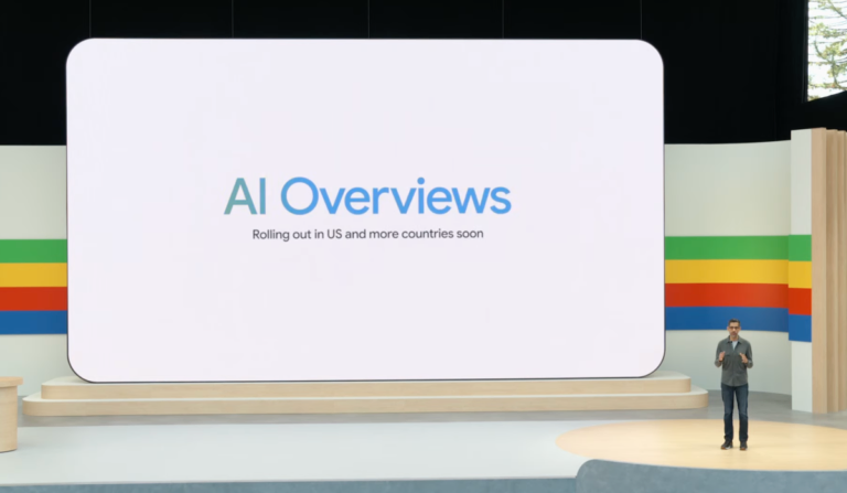 Person standing on stage beside a large screen displaying "AI Overviews, rolling out in US and more countries soon.