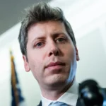 Person in a suit stands indoors, looking forward with a neutral expression. A flag is blurred in the background.