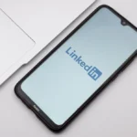Smartphone displaying LinkedIn logo on screen, placed next to a laptop on a white surface.