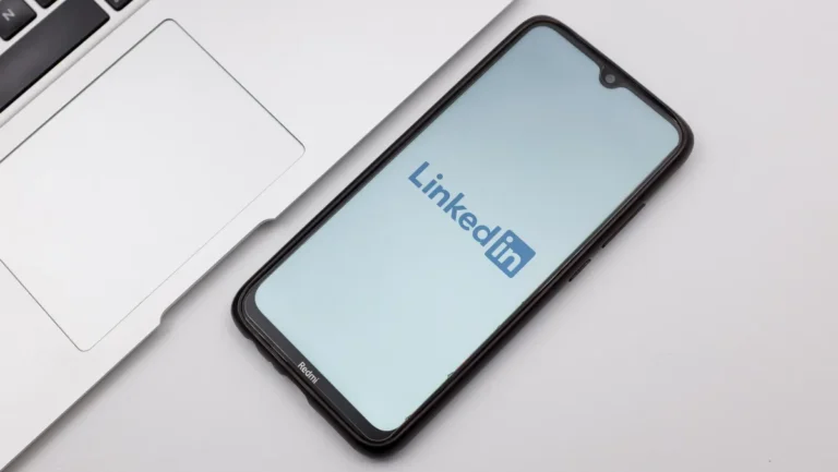 Smartphone displaying LinkedIn logo on screen, placed next to a laptop on a white surface.