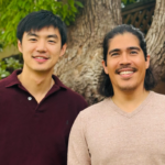 Two men smiling outdoors, standing in front of a tree and wooden fence, wearing casual shirts.