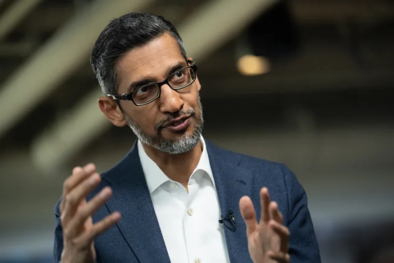 Man in glasses speaking, gesturing with hands, wearing a blue suit and white shirt, indoor setting with blurred background.