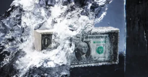 Stack of US dollar bills encased in ice, partially melting, with water splashing around.