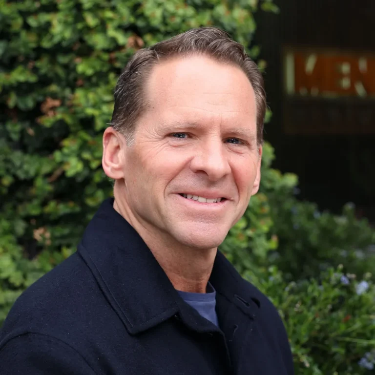 Man in a dark jacket smiling outdoors, with greenery in the background.