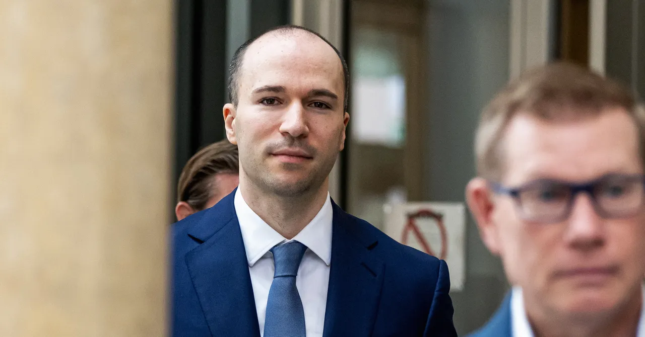 Man in a blue suit and tie standing outdoors with a blurred background, another person partially visible in the foreground.