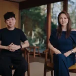 Two people seated indoors, one in a black outfit and one in blue, with large windows showing a forested view.