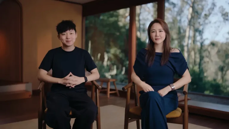 Two people seated indoors, one in a black outfit and one in blue, with large windows showing a forested view.
