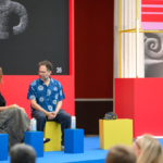 Two people seated on colorful blocks, engaging in conversation on stage with abstract art and the number 26 in the backgro...