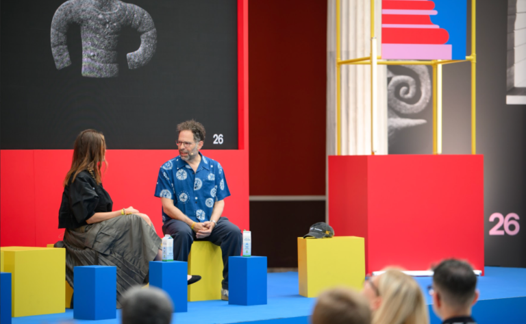 Two people seated on colorful blocks, engaging in conversation on stage with abstract art and the number 26 in the backgro...