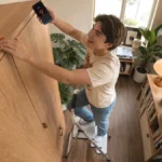 Person standing on a step stool, holding a phone with a key icon, in a cozy room with plants and bookshelves.