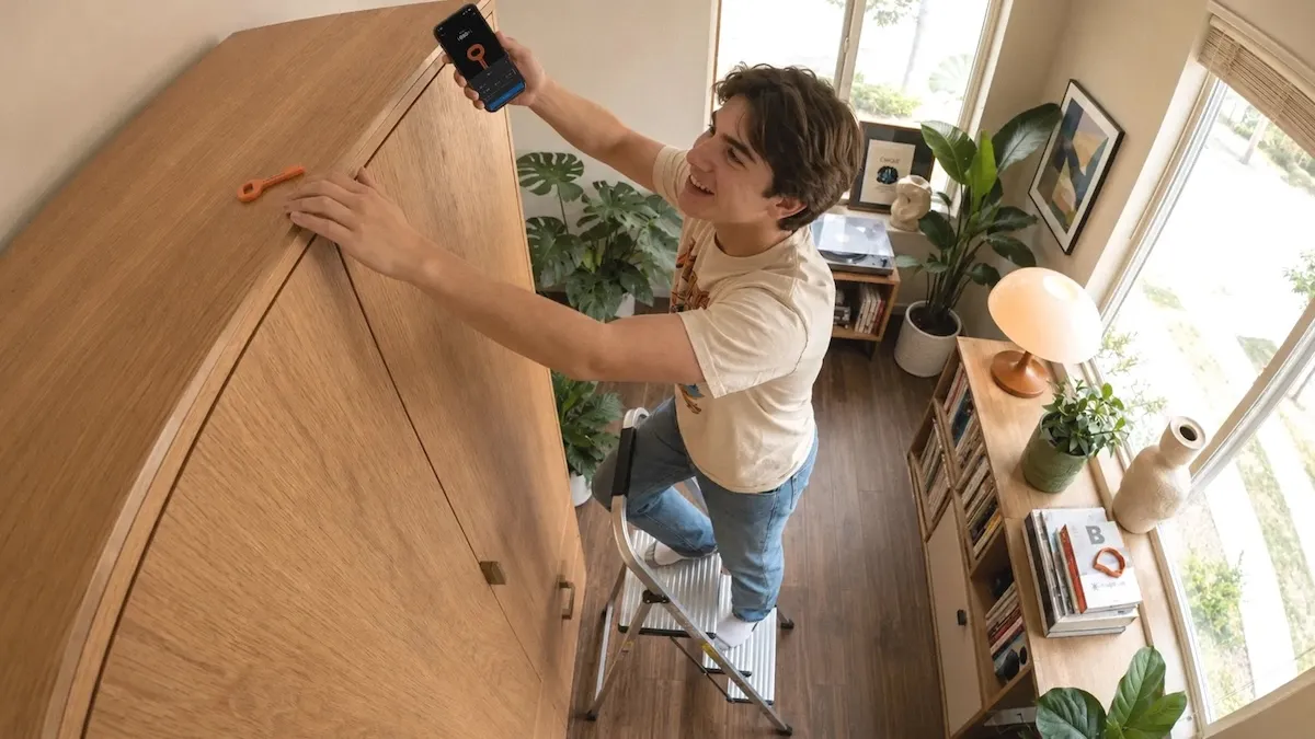 Person standing on a step stool, holding a phone with a key icon, in a cozy room with plants and bookshelves.