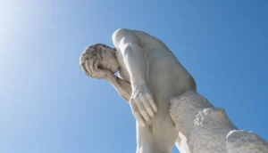 Stone statue of a person with hand covering face against a clear blue sky, viewed from below.