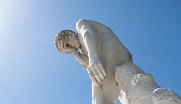Stone statue of a person with hand covering face against a clear blue sky, viewed from below.