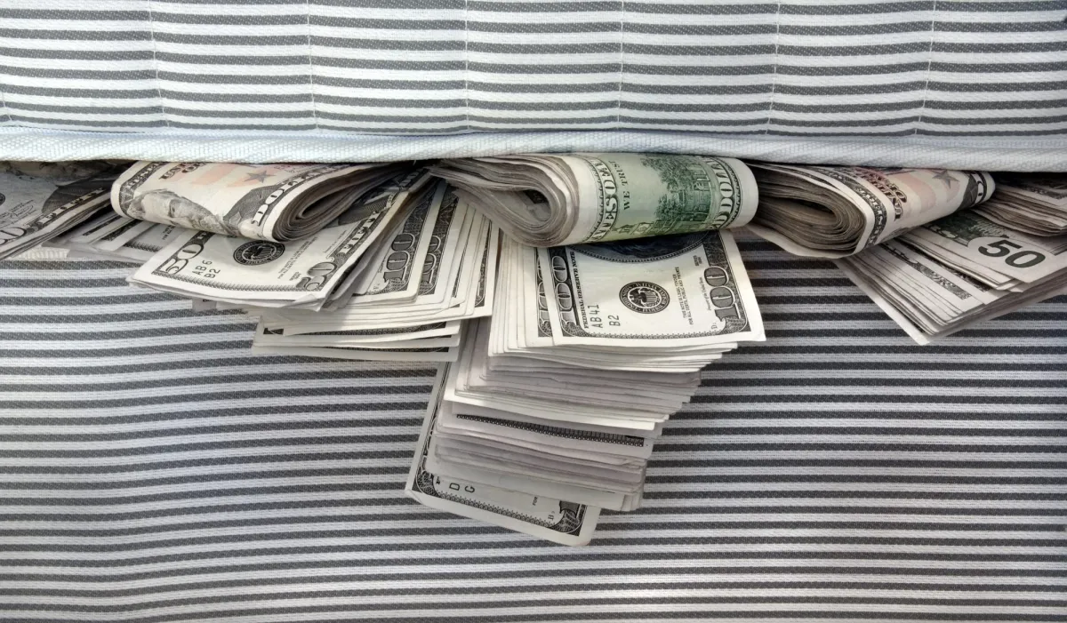 Stacks and rolls of U.S. dollar bills hidden under a striped mattress.