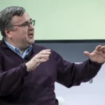 Man in a dark sweater speaking and gesturing with hands on stage, microphone clipped to shirt, blurred text in the backgro...