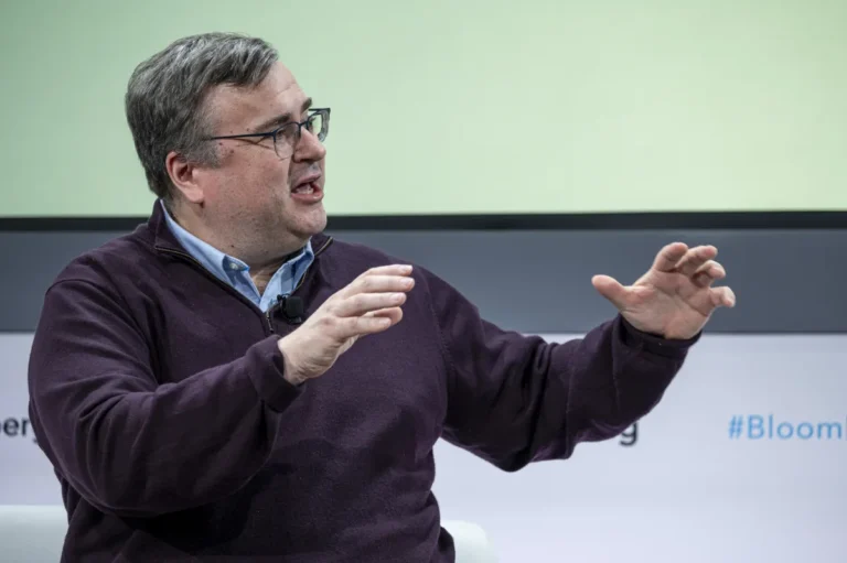 Man in a dark sweater speaking and gesturing with hands on stage, microphone clipped to shirt, blurred text in the backgro...