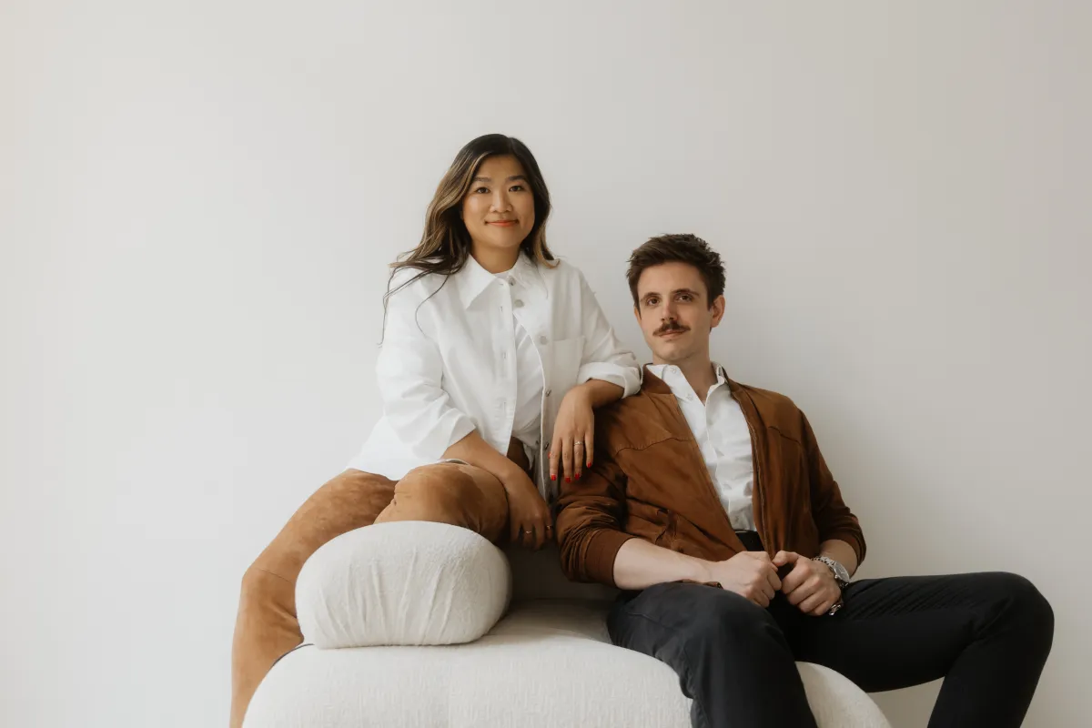 Two people sitting on a white couch against a plain background, one wearing a white shirt, the other a brown jacket.