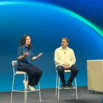 Two people seated on stage discuss in front of a blue and green backdrop, with a desk and camera in the foreground.
