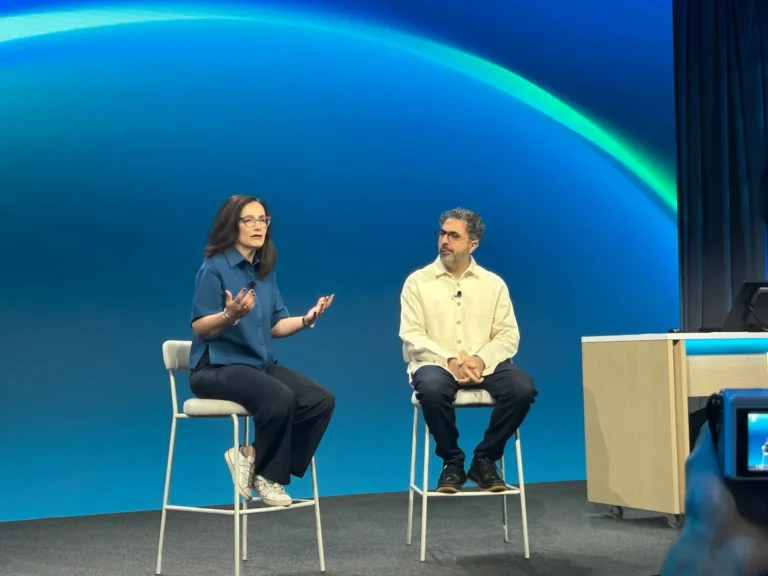 Two people seated on stage discuss in front of a blue and green backdrop, with a desk and camera in the foreground.