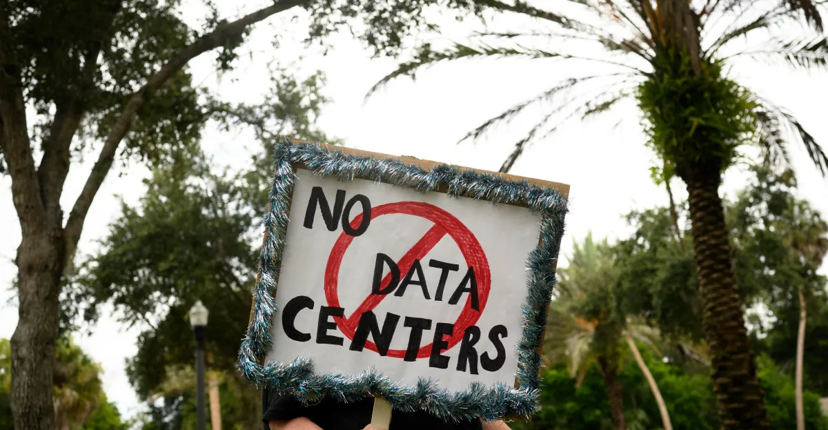 Protest sign reading "No Data Centers" with a red circle and slash, held in front of palm trees and greenery.