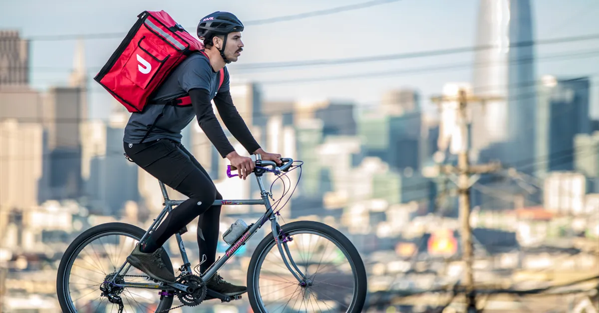 Cyclist with a red delivery backpack rides through an urban setting, wearing a helmet and black attire.