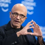 Bald man in glasses speaking, hands gesturing, blue background with blurred text "World Economic Forum".
