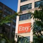 Etsy logo displayed on a large office building, partially obscured by leafy branches in the foreground.