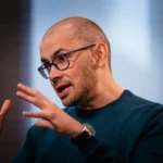 Bald man with glasses and a beard gestures while speaking, wearing a teal sweater in a softly lit indoor setting.
