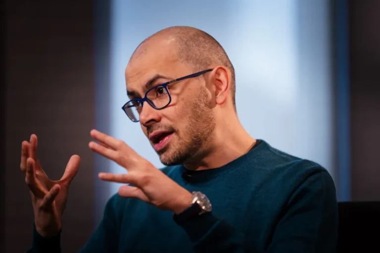 Bald man with glasses and a beard gestures while speaking, wearing a teal sweater in a softly lit indoor setting.