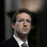 A man in a suit and tie appears serious, with a blurred dark background.