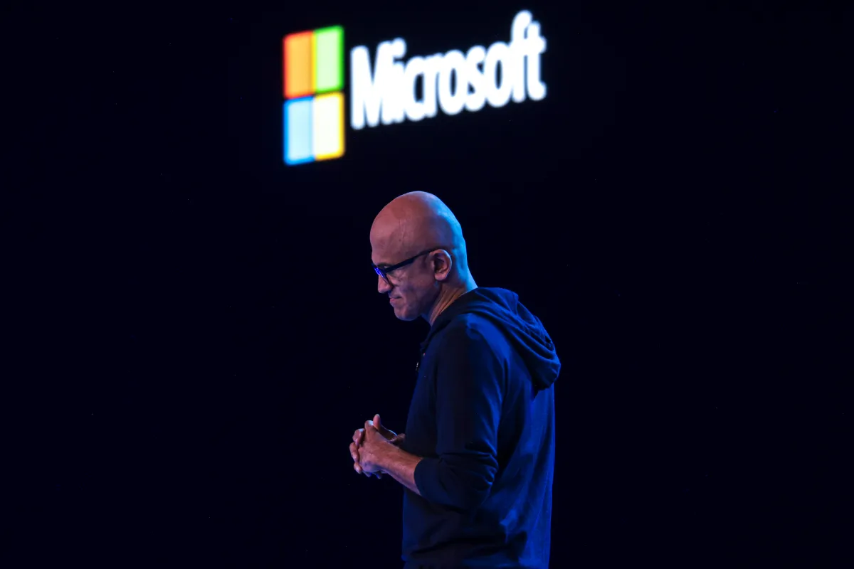 Man in dark clothing stands on stage with Microsoft logo in the background.