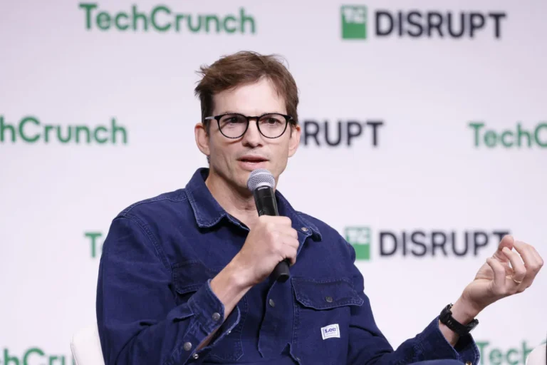 Man in glasses speaking into a microphone at TechCrunch Disrupt event, wearing a blue denim shirt.