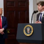 Two men in suits at a podium with the U.S. presidential seal, one speaking into a microphone. American flag in the backgro...