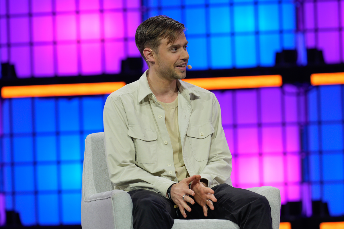 Man seated on stage, wearing a beige shirt, speaking with a microphone. Background features colorful geometric light panels.