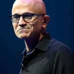 Man in black shirt with glasses standing against a dark blue background.